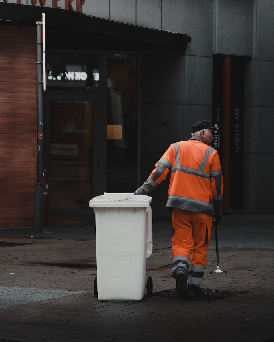 man in orange jacket and black pants standing beside white plastic trash bin